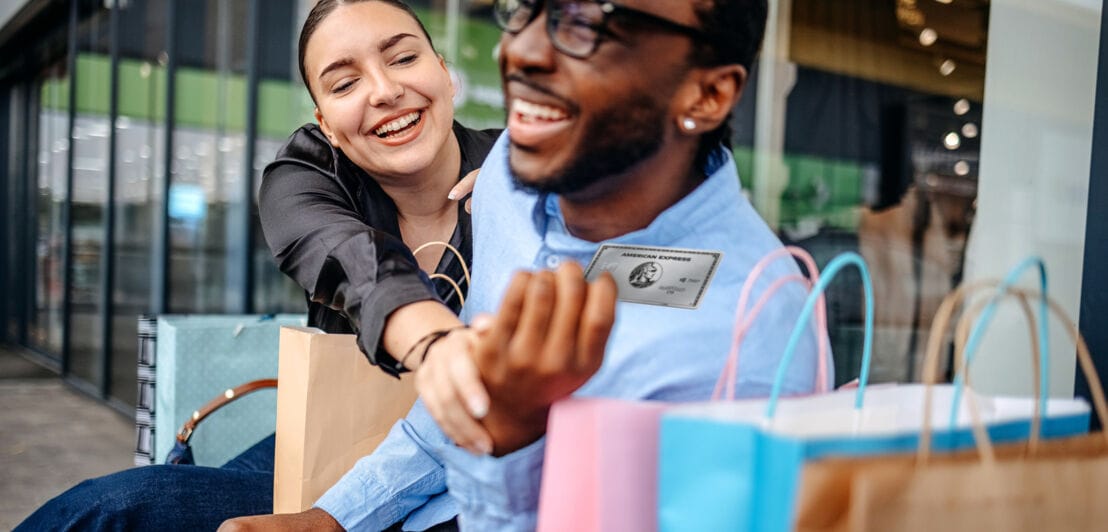 Eine Frau und ein Mann sitzen gut gelaunt neben Einkaufstüten auf einer Bank vor einem Geschäft, sie greift nach einer silbernen Kreditkarte von American Express in seiner Hand.