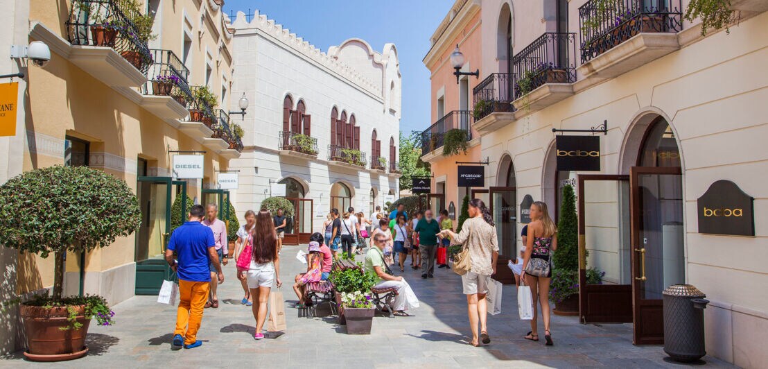 Personen laufen durch eine Fußgängerzone mit Sitzbänken vorbei an Geschäften in einem Freiluft-Einkaufszentrum bei Sonnenschein.
