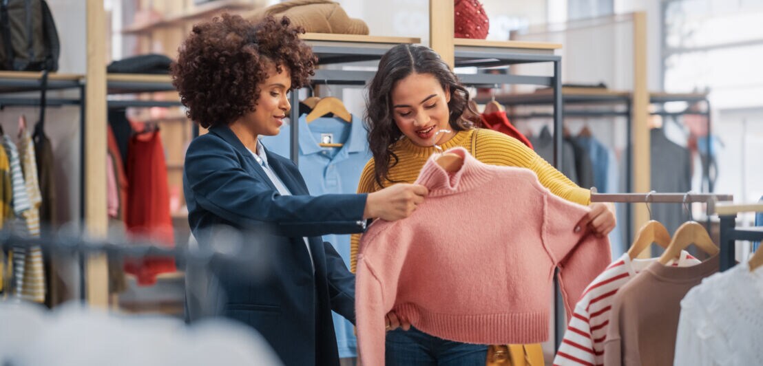 wei Frauen stehen in einem Modegeschäft und begutachten einen rosafarbenen Strickpullover in ihren Händen.