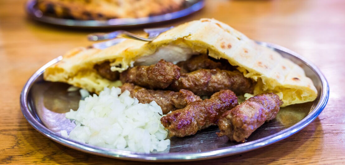 Metallteller mit mehreren gegrillten Cevapcici, gehackten Zwiebeln und Fladenbrot auf Holztisch.