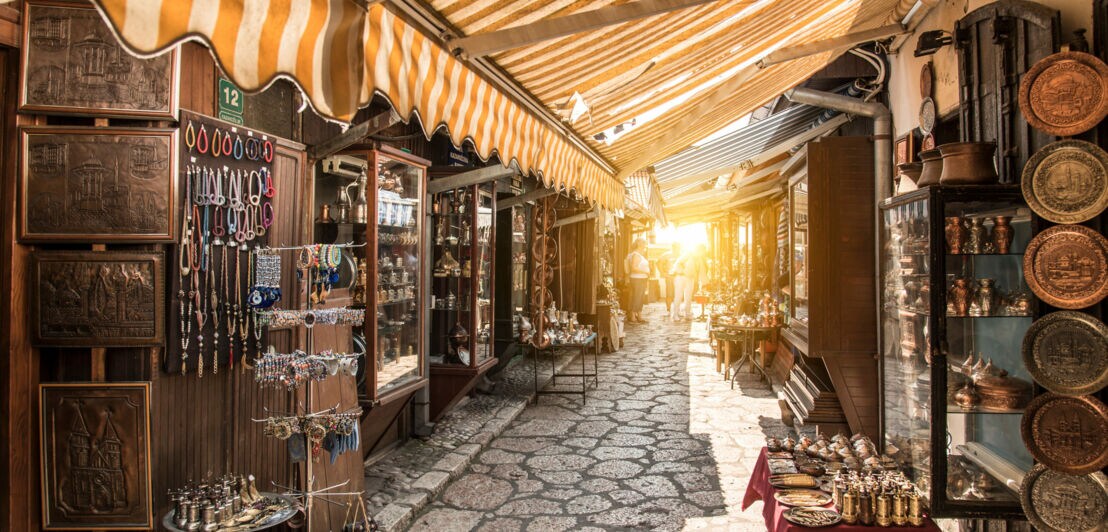 Enger Marktgang mit gepflastertem Boden, beidseitig Stände mit Schmuck, Keramik und anderen Waren unter gelb-weiß gestreiftem Markisenstoff.