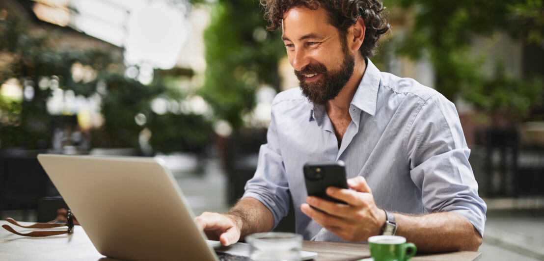 Ein Mann im legeren Hemd arbeitet an einem Laptop, neben sich eine Espressotasse, in einer Hand hält er ein Smartphone.