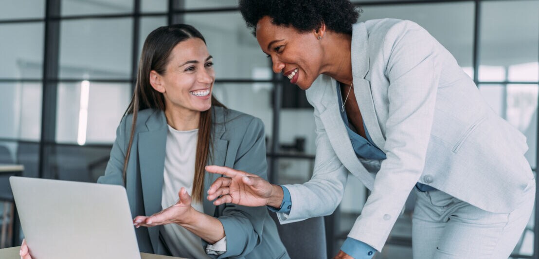 Zwei Frauen in grauen Blazern kommunizieren lächelnd an einem Arbeitsplatz mit Laptop.