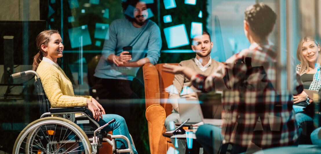 Eine Frau im Rollstuhl und weitere Personen reden in einem Raum mit Glaswand.