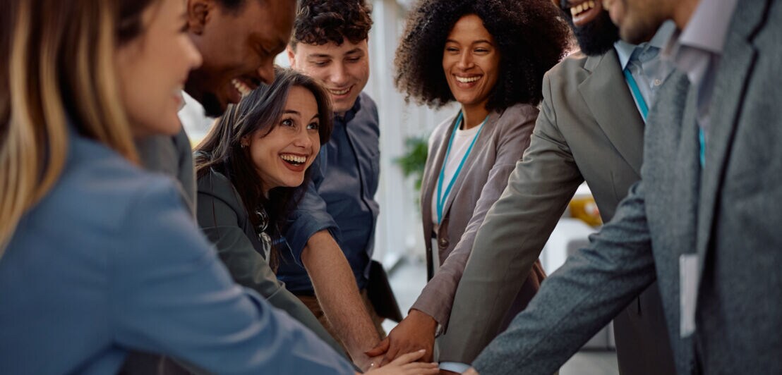Sieben lächelnde Personen in Business-Kleidung stehen im Kreis und legen die Hände übereinander.