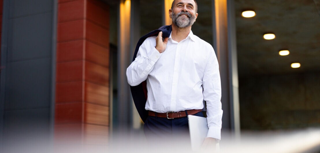 Ein lächelnder Mann im weißen Hemd steht mit einem über die Schulter geworfenen Jackett und Laptop in der Hand vor einem Gebäude.