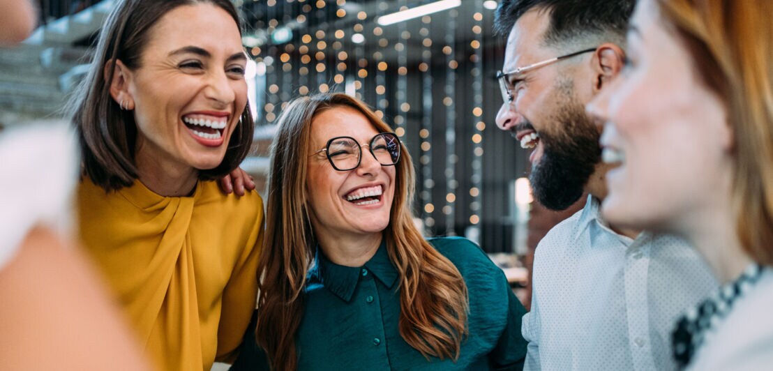 Vier lachende Personen in eleganter Kleidung stehen eng beieinander in einem Büro.