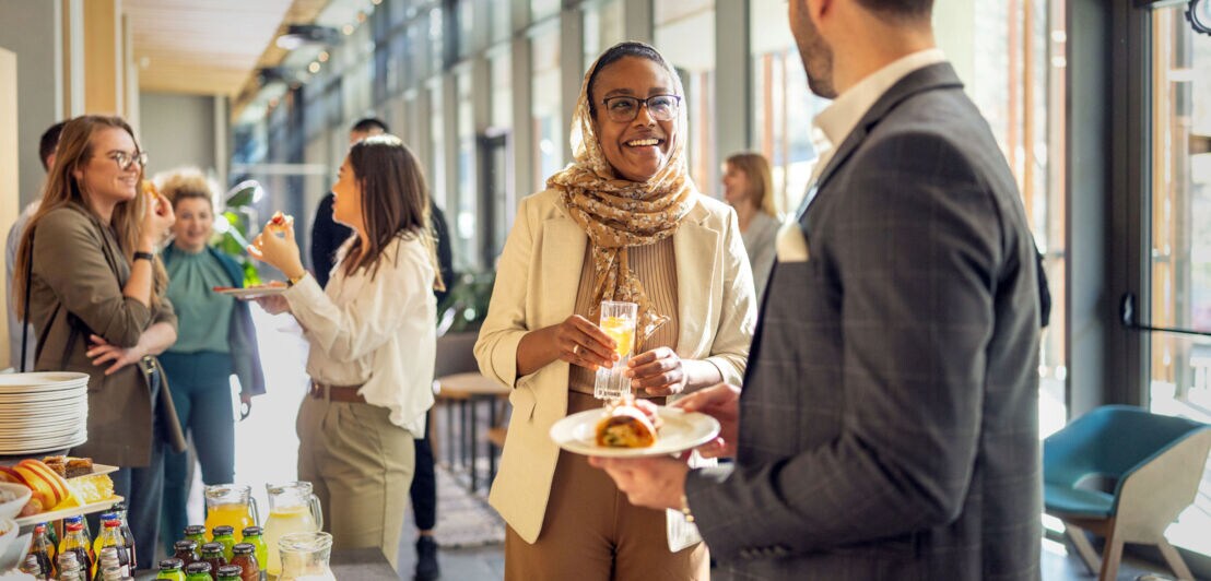 Eine Frau und ein Mann unterhalten sich an einem Büfett in einem hellen Foyer, im Hintergrund stehen weitere Personen.