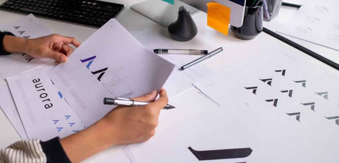 Entwurfsgestaltung eines Logos auf Papierblättern in den Händen einer Person auf einem Schreibtisch.