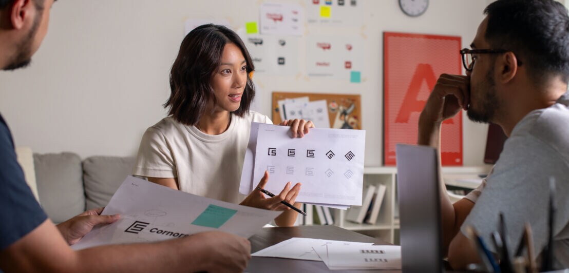 Eine Frau sitzt an einem Tisch zwischen zwei weiteren Personen und präsentiert Logoentwürfe auf einem Papierblatt in ihren Händen in einem Arbeitszimmer.