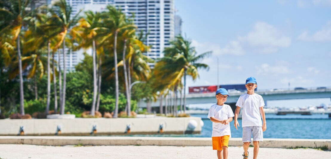 Zwei Jungen mit Schirmmützen spazieren an der Küste Miamis, im Hintergrund Palmen, Hochhäuser und das Meer.