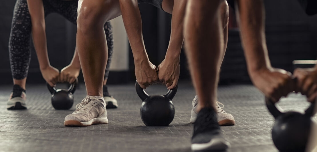Drei Personen in Sportkleidung halten Kettlebells mit beiden Händen, Blick auf die Beine und Hände, Fitnessstudio-Boden sichtbar.