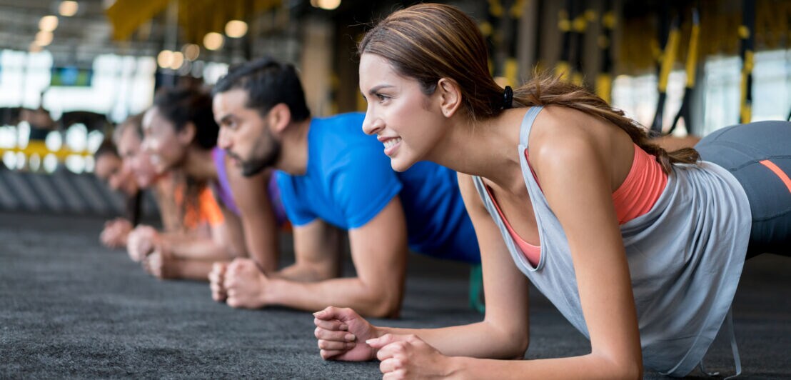 Gruppe von Menschen in Sportkleidung macht Plank-Übung auf Boden in Fitnessstudio.
