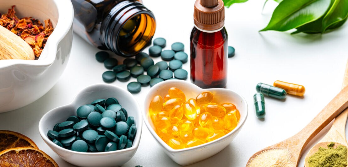 Herzförmige Schalen mit grünen Tabletten und gelben Kapseln, umgeben von weiteren Kapseln, getrockneten Orangenscheiben, zwei Holzlöffeln mit Pulver und grünen Blättern.