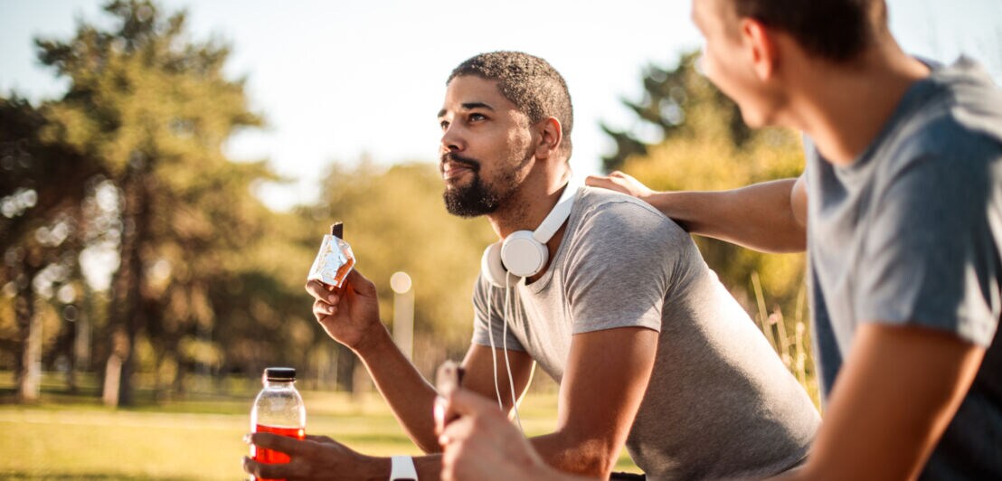 Zwei Männer sitzen draußen auf einer Bank, einer hält eine Wasserflasche und einen Proteinriegel, der andere legt die Hand auf dessen Schulter.