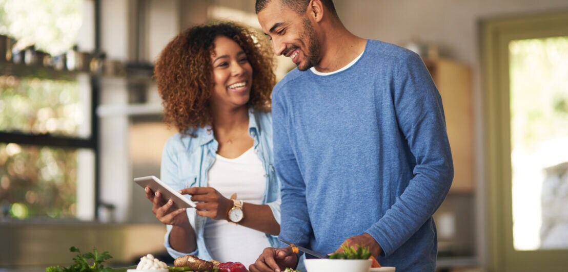 Zwei Personen stehen in einer Küche, eine hält ein Tablet, auf dem Tisch liegen verschiedene Schüsseln mit Lebensmitteln.