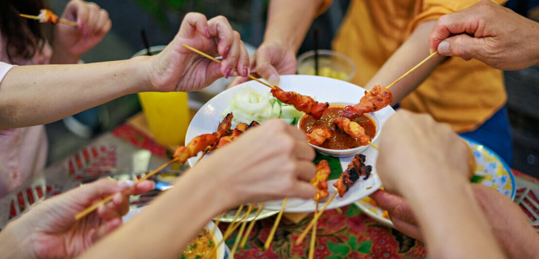 Mehrere Hände dippen Hühnchenspieße in eine braune Soße auf einem Teller in ihrer Mitte an einem Tisch.