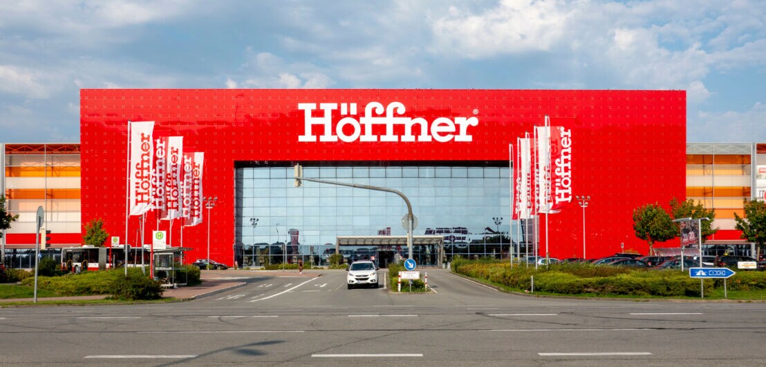 Großes, freistehendes Gebäude mit Schriftzug „Höffner“ auf roter Fassade hinter einer Straßenkreuzung mit rot-weißen Fahnen.