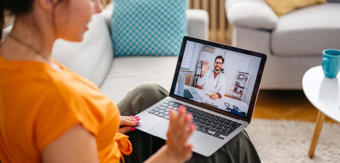 Eine Frau sitzt auf einem Sofa mit einem Laptop auf den Beinen und kommuniziert via Videobesprechung mit einem Arzt.