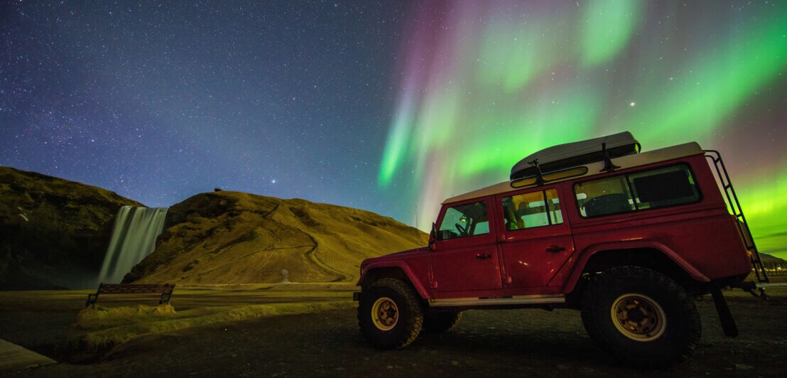 Ein roter Jeep parkt in einem Naturgebiet mit Wasserfall bei Polarlichtern am nächtlichen Sternenhimmel.
