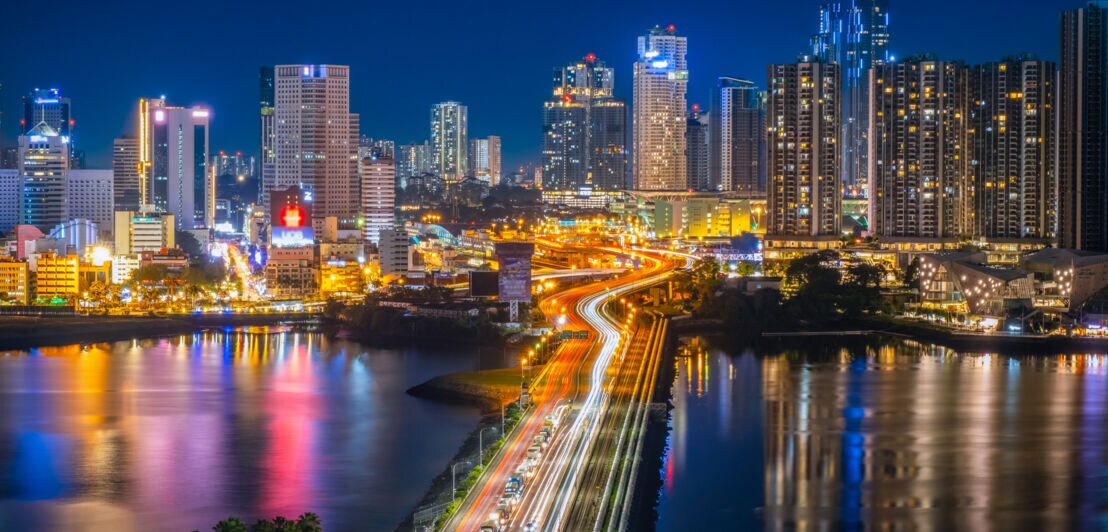 Nächtliche Stadtansicht mit beleuchteten Hochhäusern und einer vielbefahrenen Brücke über Wasser, Lichtspuren von Fahrzeugen sichtbar.