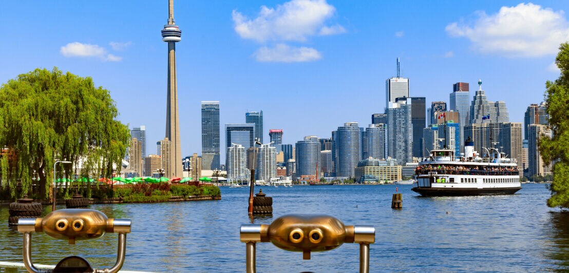 Skyline von Toronto am See mit Fähre zwischen begrünten Ufern, im Vordergrund zwei Aussichtsfernrohre aus Metall.
