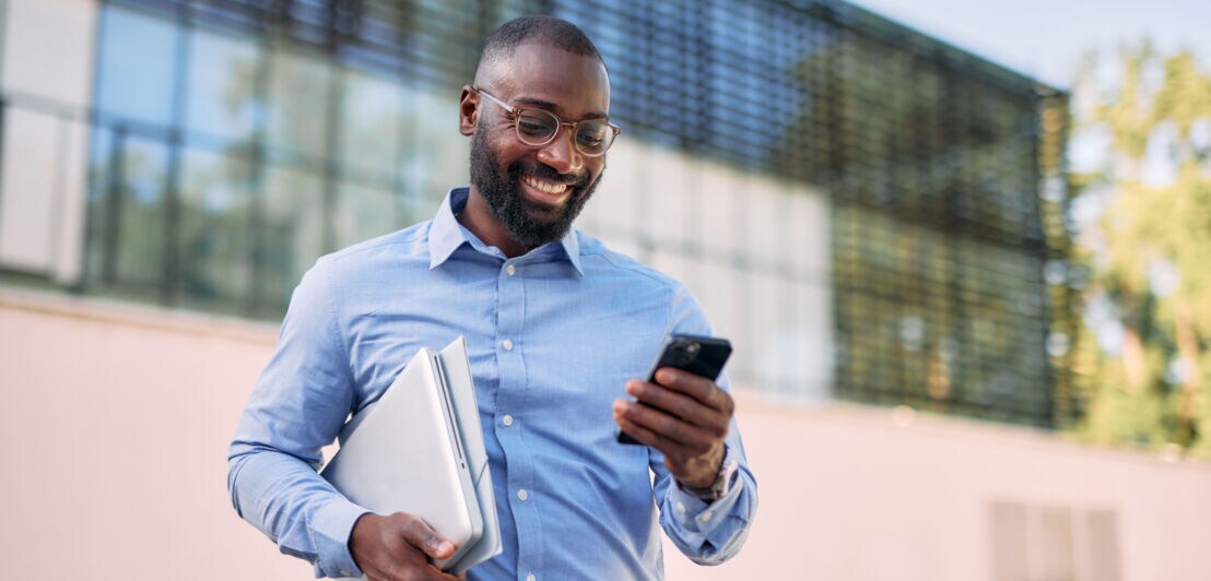 Ein Mann im blauen Hemd mit Laptop unter dem Arm steht vor einem Bürogebäude und blickt erfreut auf ein Smartphone in seiner Hand.