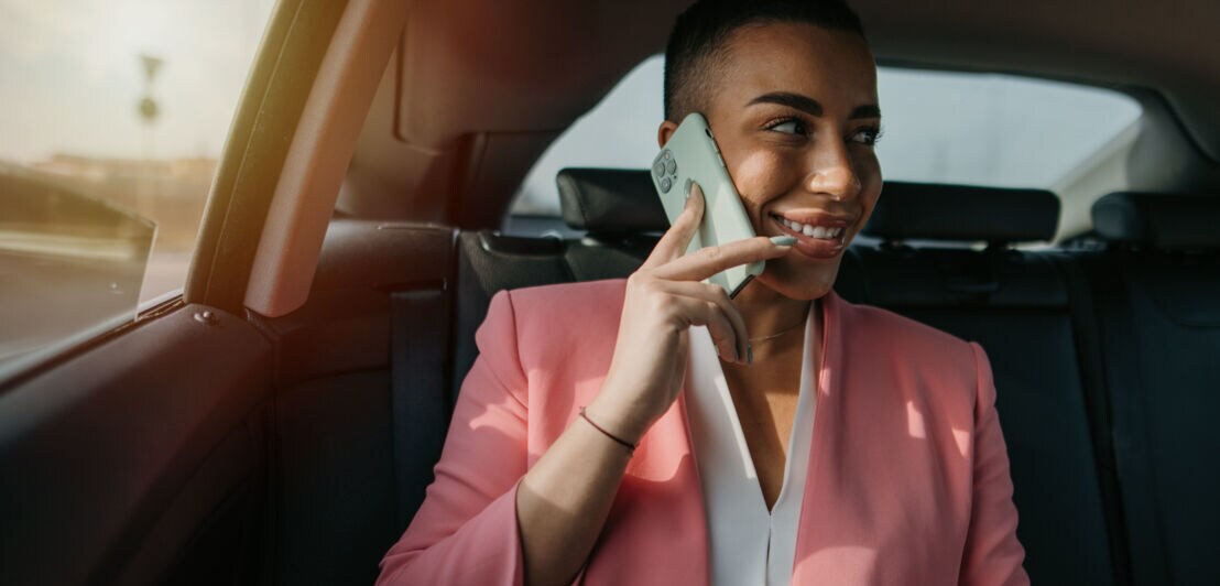 Eine lächelnde Frau im rosafarbenen Blazer sitzt auf der Rückbank eines Autos und telefoniert mit einem Smartphone an ihrem Ohr.