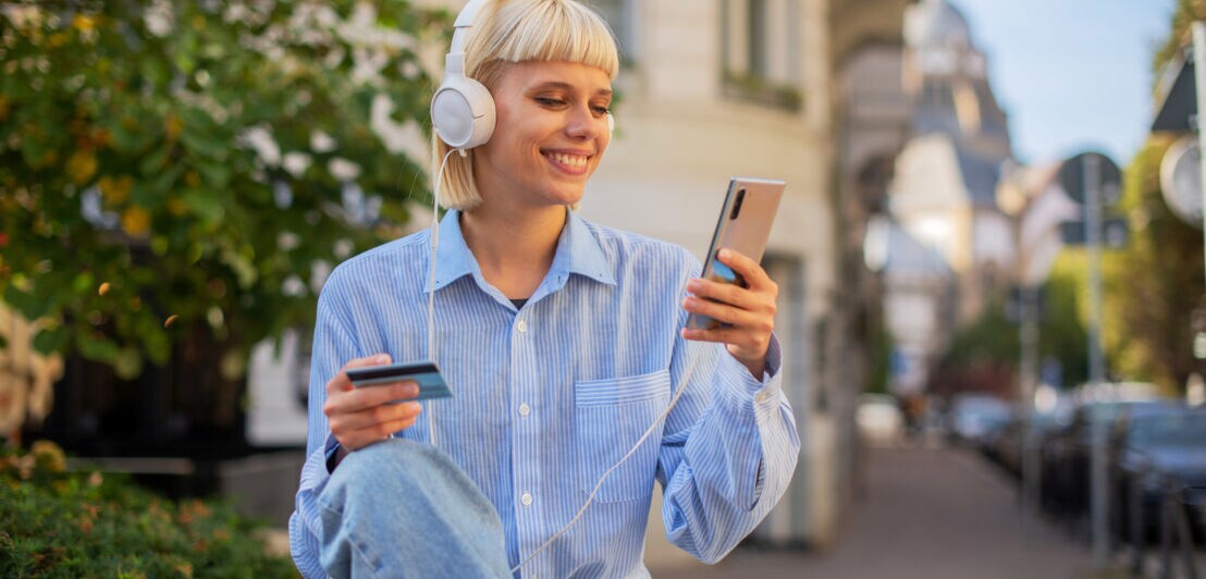 Eine junge Frau mit Kopfhören sitzt in urbaner Umgebung und blickt lächelnd auf ein Smartphone in ihrer Hand, in der anderen Hand hält sie eine Kreditkarte.
