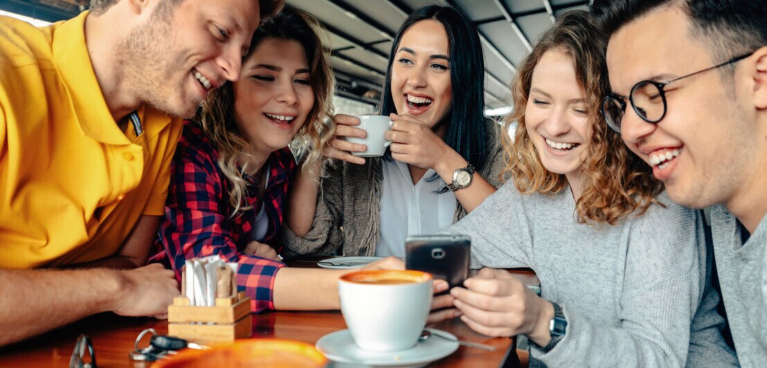 Fünf junge Erwachsene sitzen gut gelaunt an einem Tisch mit Kaffeetassen und schauen gemeinsam auf ein Smartphone.