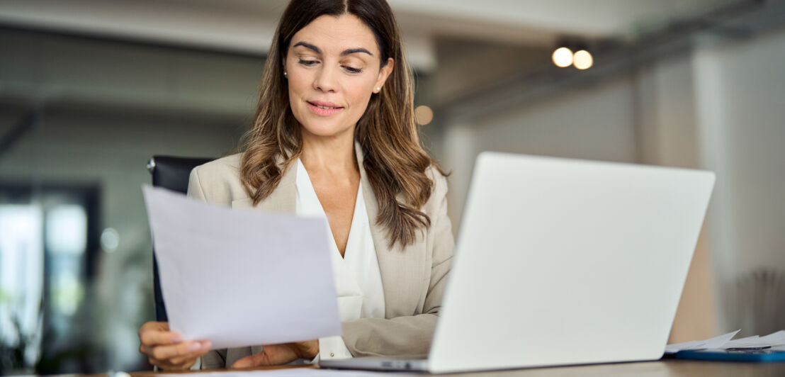 Eine Frau in Business-Kleidung sitzt an einem Schreibtisch an einem Laptop und schaut auf ein Papierblatt in ihrer Hand.