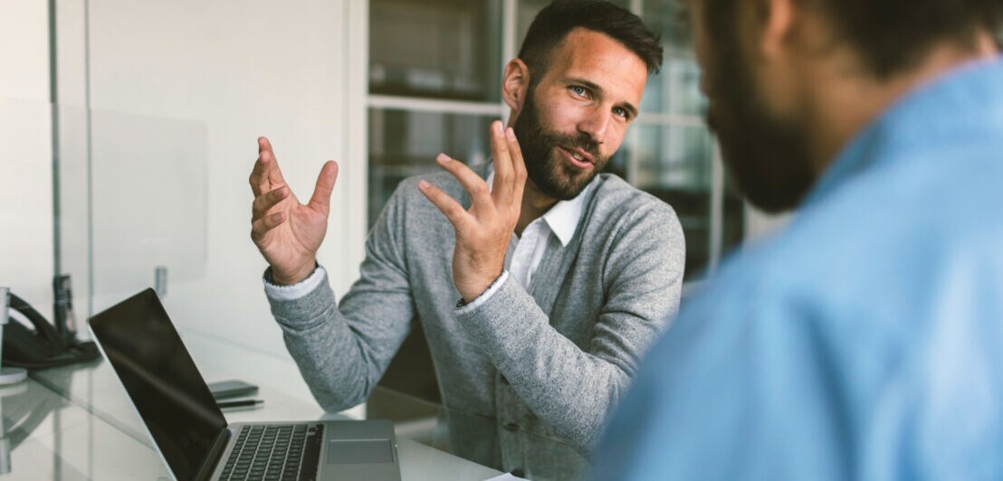 Ein Mann gestikuliert mit den Händen während eines Gesprächs mit einer Person an einem Tisch mit Laptop.