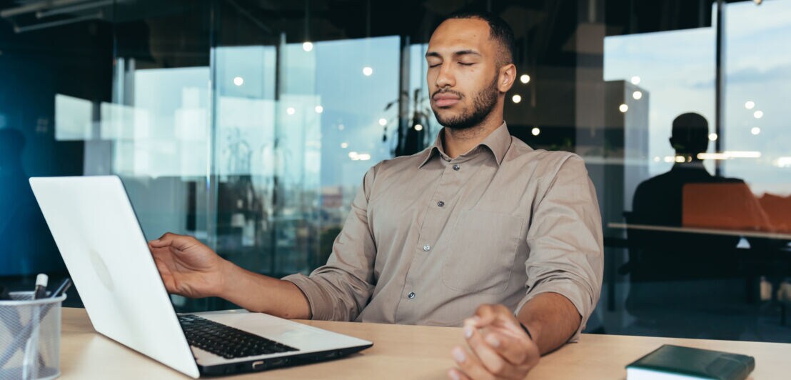 Ein Mann sitzt mit geschlossenen Augen in Meditationshaltung vor einem Laptop an einem Schreibtisch in einem Büro.