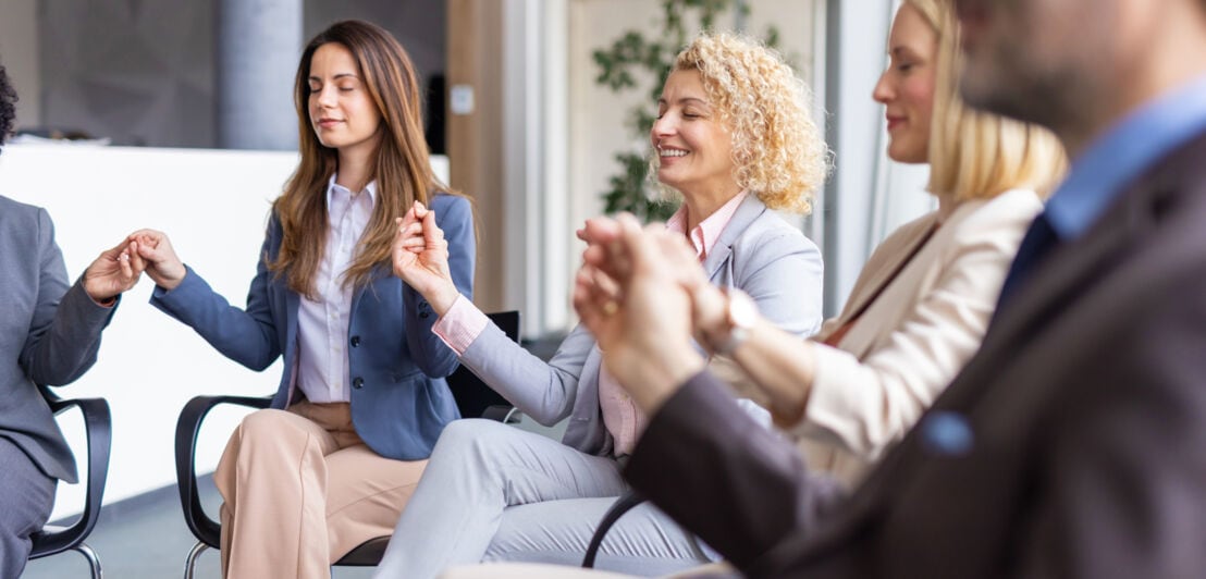 Mehrere Personen in Business-Kleidung sitzen mit geschlossenen Augen in einem Stuhlkreis in einem Büro und fassen sich an den Händen an.