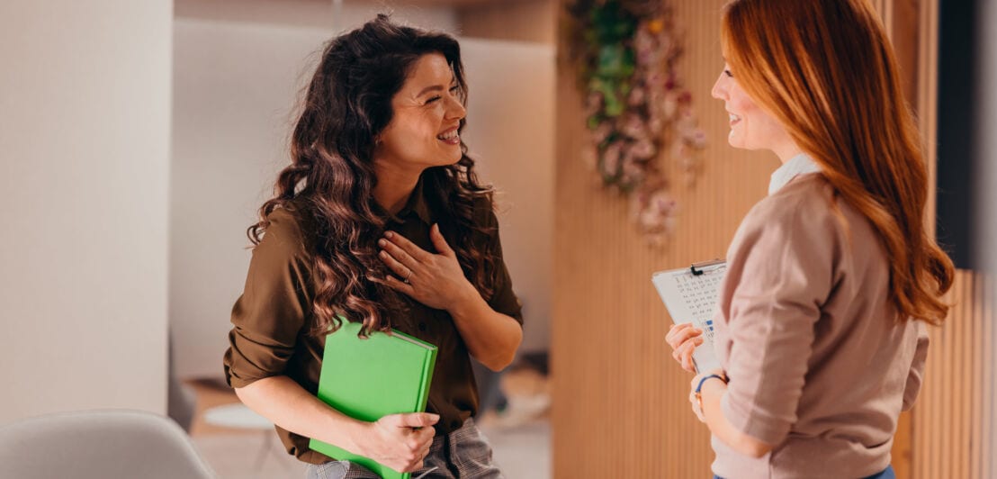 Zwei lächelnde Frauen während einer herzlichen Unterhaltung in einem Konferenzraum an, eine Frau legt dabei ihre Hand aufs Herz.