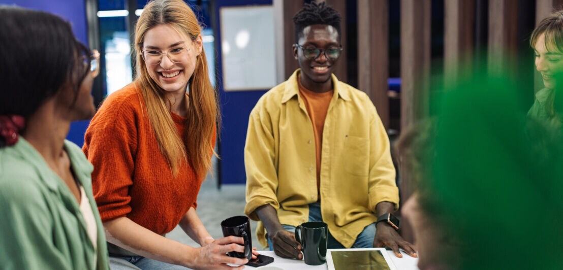 Eine Gruppe junger Menschen sitzt lächelnd beieinander, zwischen ihnen Kaffeetassen und Tablets auf einem Tisch.