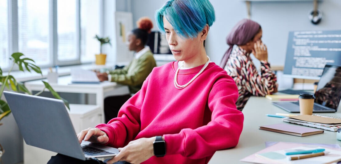 Ein junger Mann mit blauen Haaren im pinkfarbenen Pullover sitzt mit Laptop auf dem Schoß in einem Büro, im Hintergrund zwei Frauen an weiteren Arbeitsplätzen.
