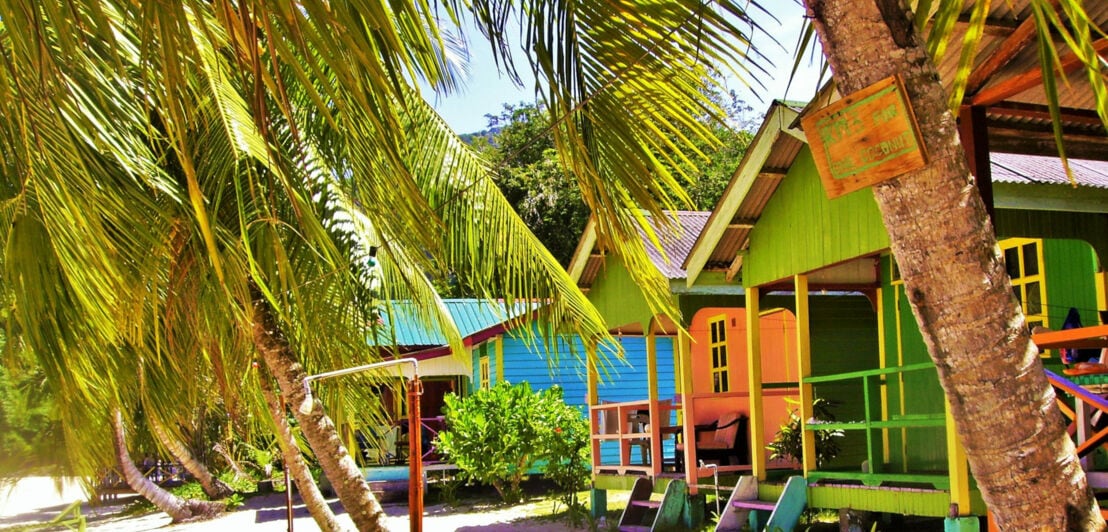 Mehrere bunte Holzhäuser auf Stelzen am Sandstrand, umgeben von Palmen und tropischer Vegetation.