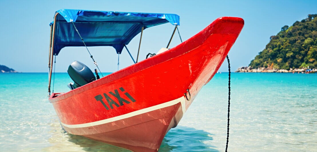 Roter Bootsrumpf mit schwarzer Aufschrift 'TAXI' am Bug, im flachen Wasser vor bewaldetem Ufer und Strand mit Palmen.