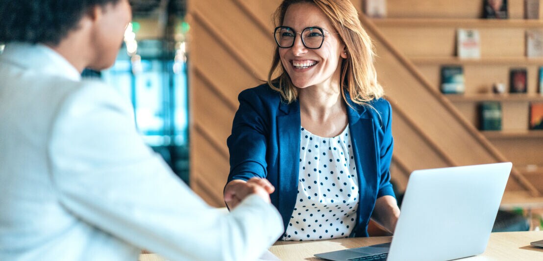 Eine lachende Frau im blauen Blazer gibt einer Person die Hand an einem Tisch, auf dem ein Laptop steht.