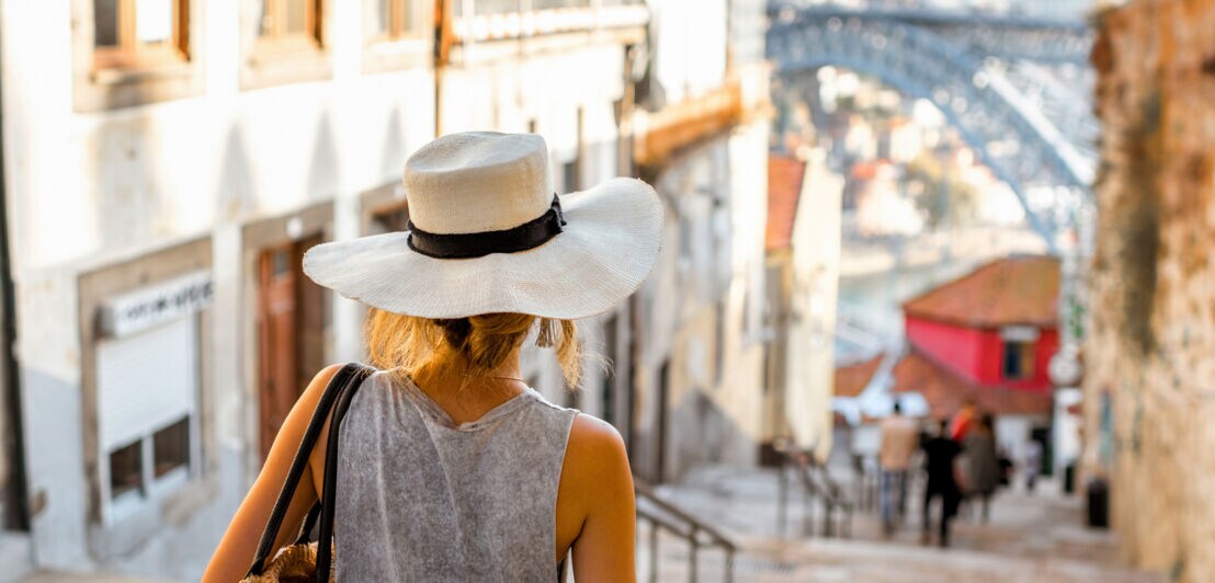 Rückansicht einer Frau mit weißem Sonnenhut, die eine Steintreppe in einer Altstadt hinuntergeht.