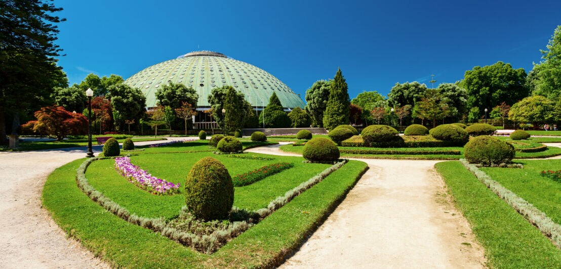 Kuppelgebäude hinter einer gepflegten Parkanlage mit Kieswegen unter blauem Himmel.