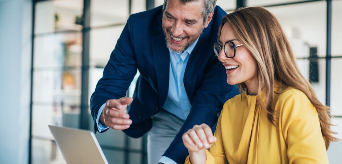Eine Frau sitzt an einem Schreibtisch, neben ihr steht ein Mann im blauen Sakko, gemeinsam schauen sie lächelnd auf ein Laptop in einem Büroraum.