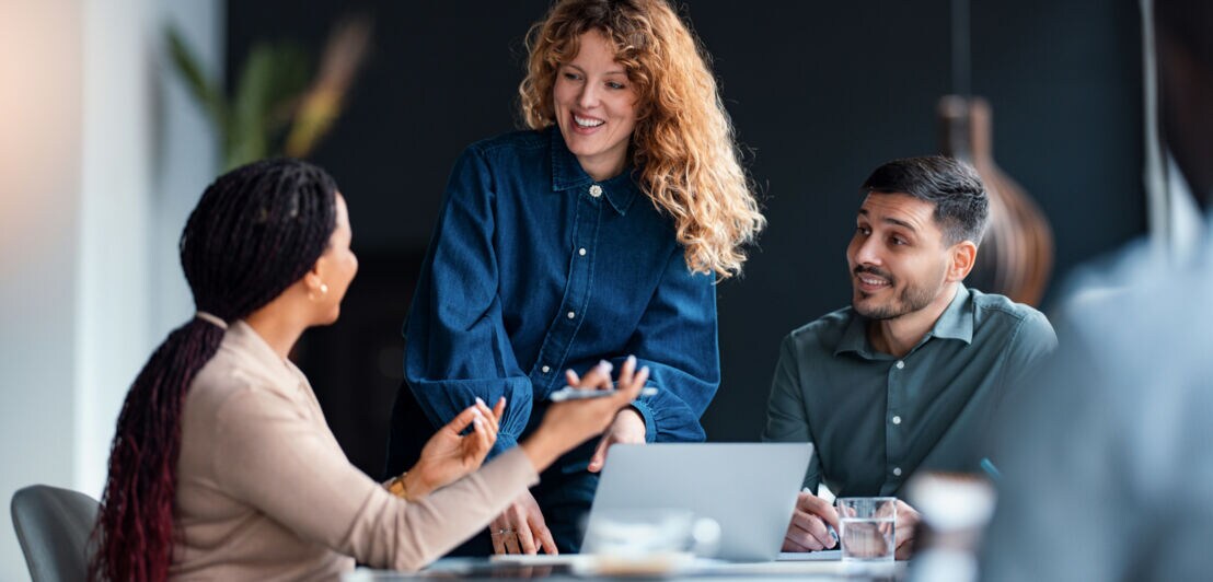 Eine Frau beugt sich über einen Tisch, auf dem ein Laptop steht und an dem zwei weitere Personen sitzen.