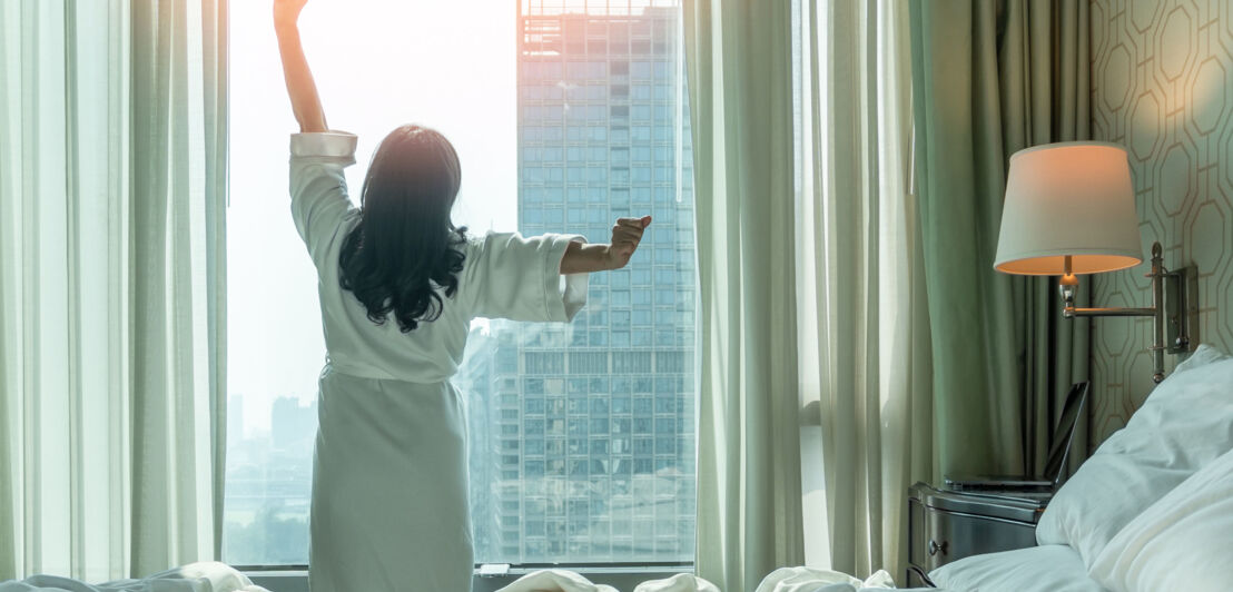 Rückansicht einer sich räkelnden Person im weißen Bademantel am Fenster eines Schlafzimmers in einer Großstadt, im Vordergrund ein Bett.