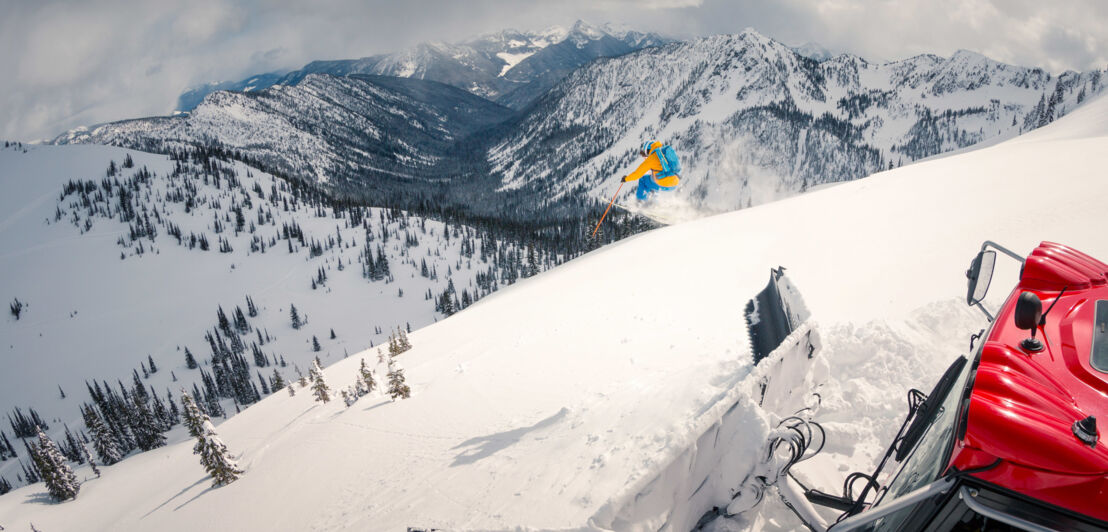 Ein Skifahrer springt einen Schneehang hinunter vor Bergpanorama, im Vordergrund eine Pistenplanierraupe im Tiefschnee.