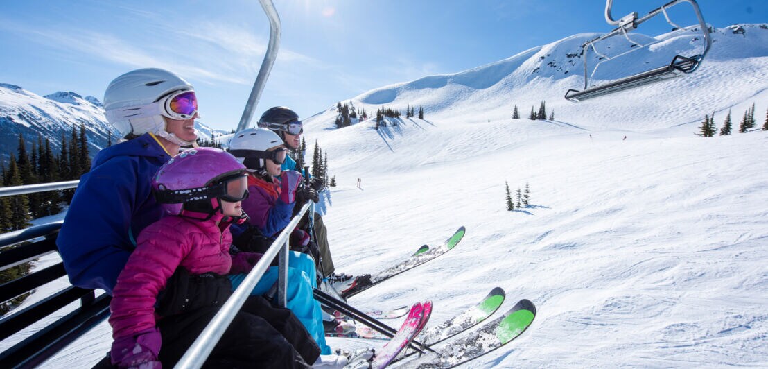 Eine Familie mit zwei Kindern auf Skiern sitzt in einem Sessellift über einer Skipiste bei Sonnenschein.