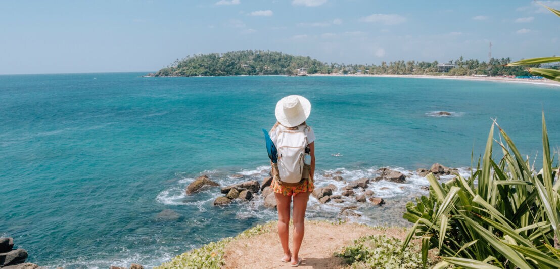 Eine Person mit Sonnenhut steht auf einer Klippe Richtung Meer, vor ihr die Küstenlandschaft Sri Lankas.
