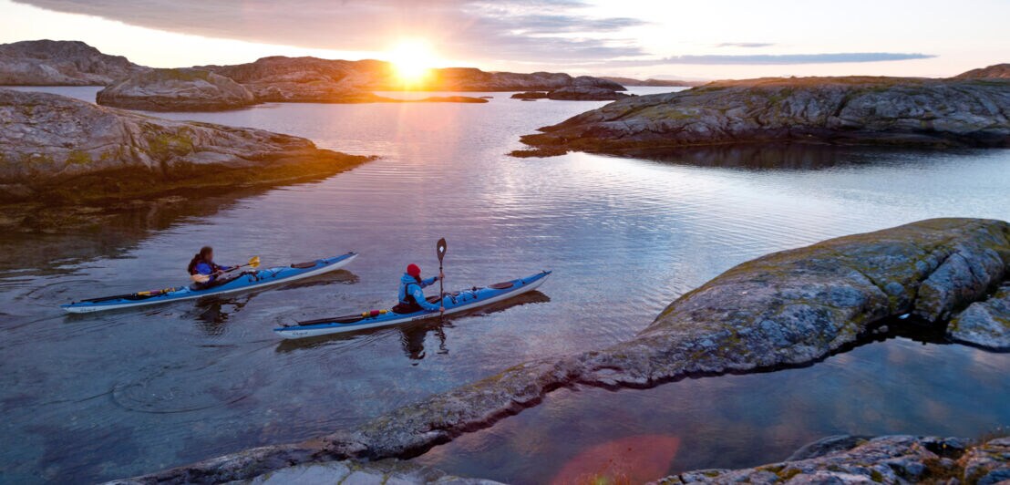 Zwei Personen paddeln in blauen Kajaks auf ruhigem Wasser zwischen felsigen Inseln bei Sonnenuntergang.