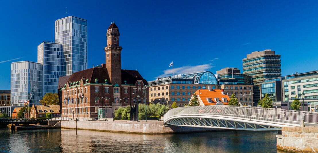 Blick auf einen Fluss mit einer modernen Fußgängerbrücke und historischen sowie modernen Gebäuden im Hintergrund unter blauem Himmel.